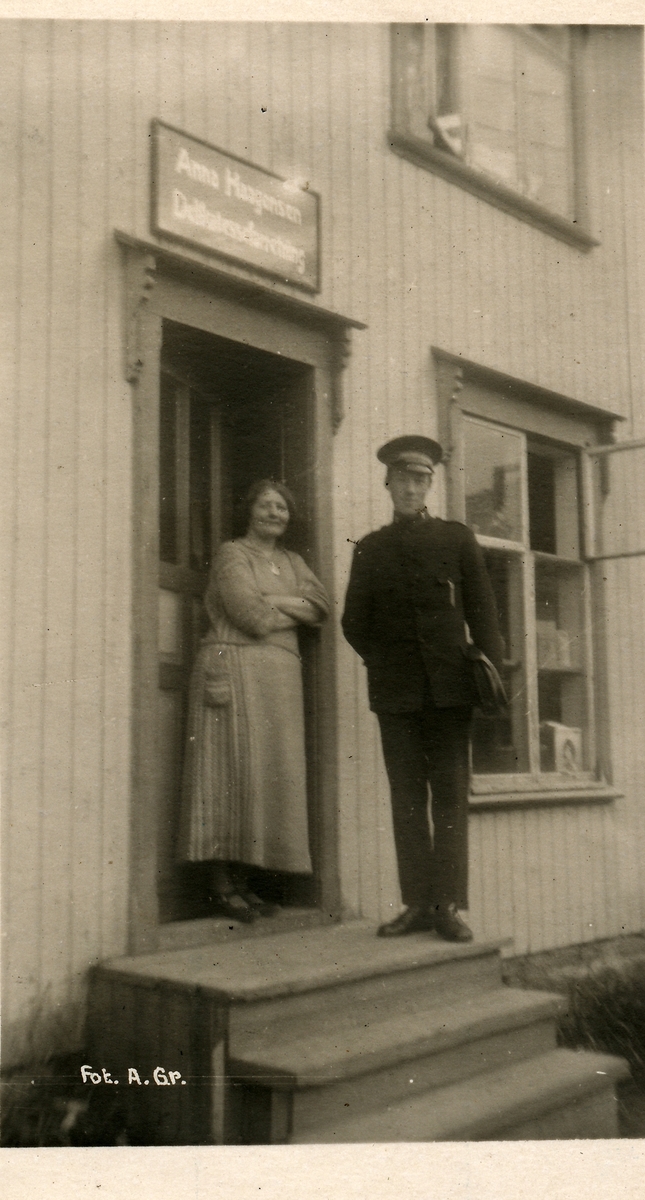Kvinne og ung mann i uniform på trapp foran inngangsdør til forretning. Over døra er det skiltet: Anna Haagensen, delikatesseforretning.