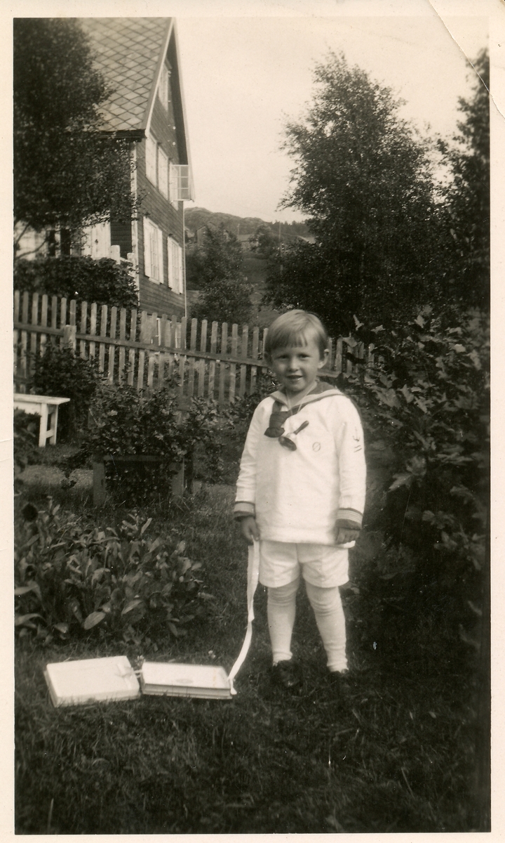 Liten gutt på 3-4 år. Han har matrosdress, bjelle og drar i noen esker. Han står i en hage med ripsbusk. Øistein Andersen.