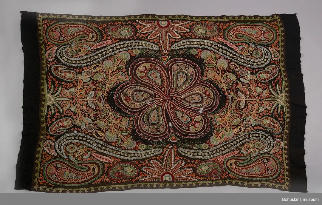 Överkast i mörk stickad (rätstickning) och virkad botten i ullgarn. Rikt broderad yta i kulörta färger (ullgarn färgat med anilinfärger).