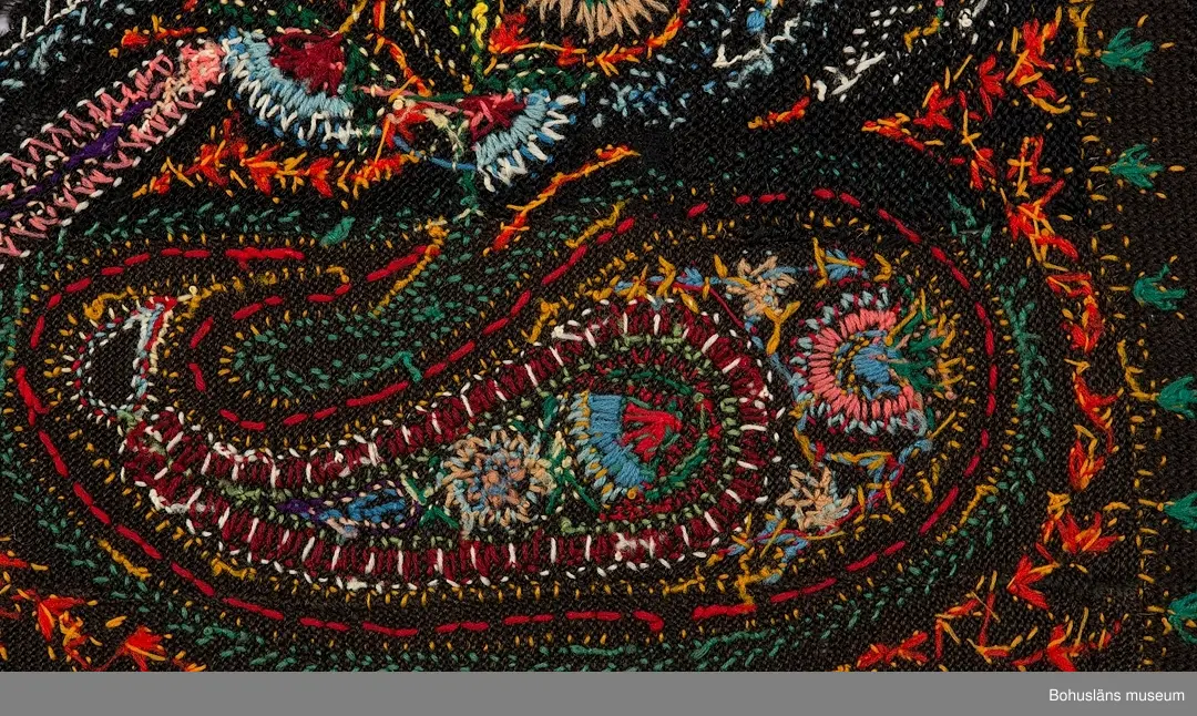 Överkast i mörk stickad (rätstickning) och virkad botten i ullgarn. Rikt broderad yta i kulörta färger (ullgarn färgat med anilinfärger).