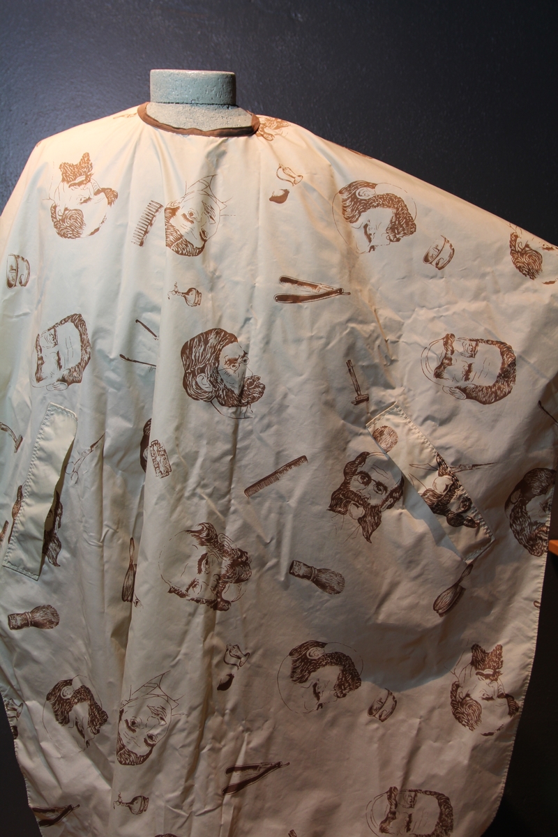 Form: avrundet, rektangulært stykke med halsringning, lukking bak. Stoffet har dekor som viser tegninger av barbersaker, børster og historiske menn med frisert hår, skjegg og/eller bart.