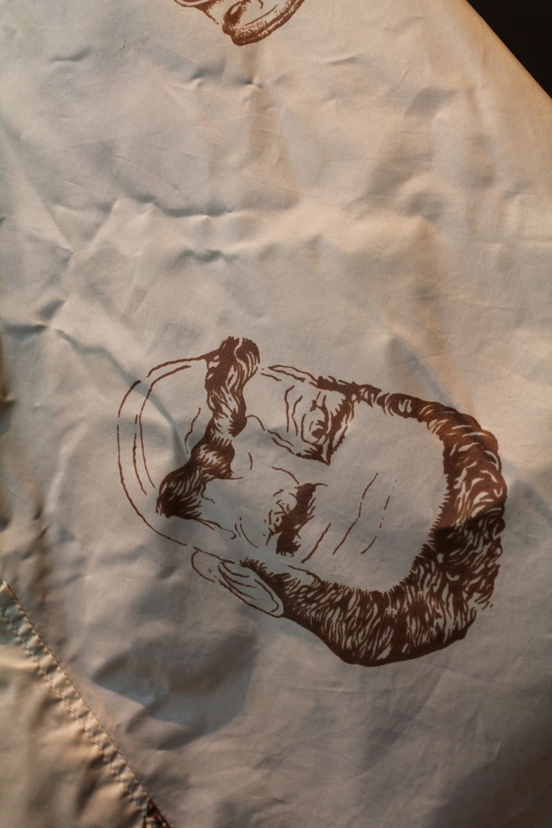 Form: avrundet, rektangulært stykke med halsringning, lukking bak. Stoffet har dekor som viser tegninger av barbersaker, børster og historiske menn med frisert hår, skjegg og/eller bart.