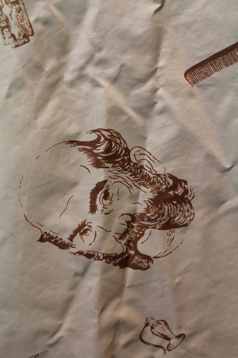 Form: avrundet, rektangulært stykke med halsringning, lukking bak. Stoffet har dekor som viser tegninger av barbersaker, børster og historiske menn med frisert hår, skjegg og/eller bart.