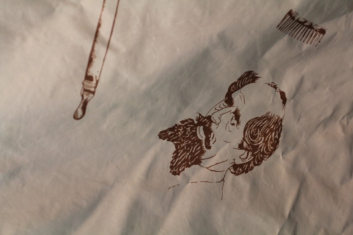 Form: avrundet, rektangulært stykke med halsringning, lukking bak. Stoffet har dekor som viser tegninger av barbersaker, børster og historiske menn med frisert hår, skjegg og/eller bart.