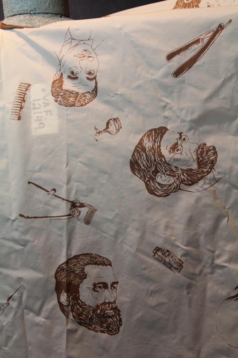 Form: avrundet, rektangulært stykke med halsringning, lukking bak. Stoffet har dekor som viser tegninger av barbersaker, børster og historiske menn med frisert hår, skjegg og/eller bart.