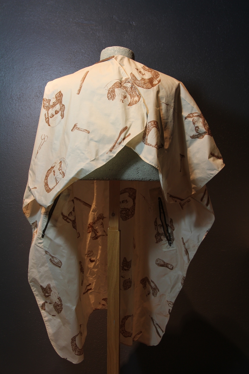 Form: avrundet, rektangulært stykke med halsringning, lukking bak. Stoffet har dekor som viser tegninger av barbersaker, børster og historiske menn med frisert hår, skjegg og/eller bart.