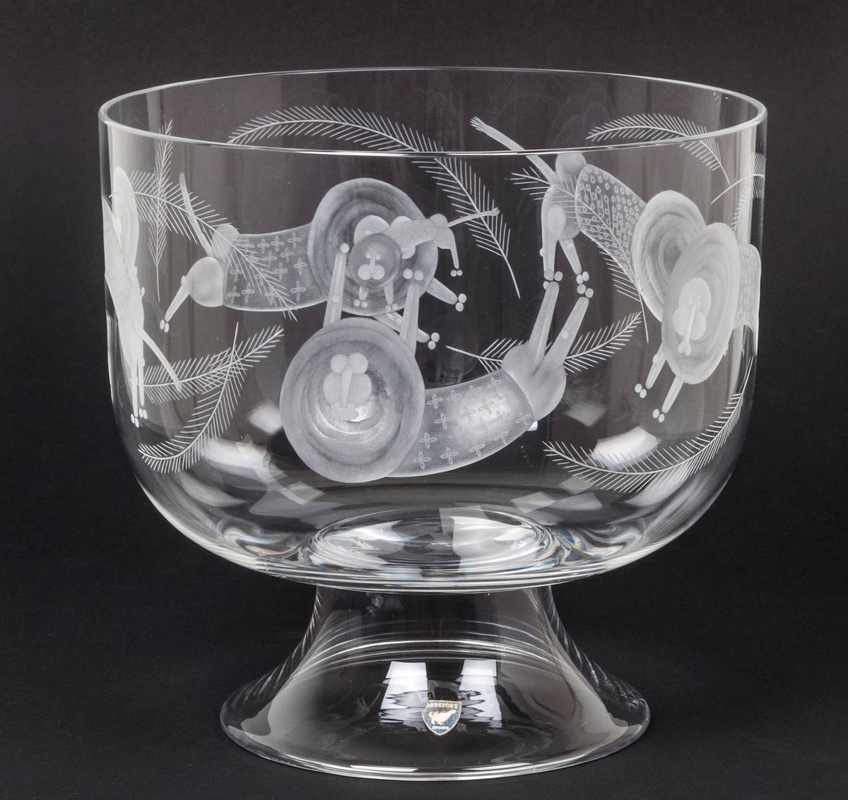 Skål på fot, kristall. Orrefors glasbruk, desing Gunnar Cyrén. Cylindrisk med välvd botten. Konkav fot. Graverad dekor av lejon och apor. På foten grå sköldformad etikett med i vitt orre och texten : ORREFORS SWEDEN.