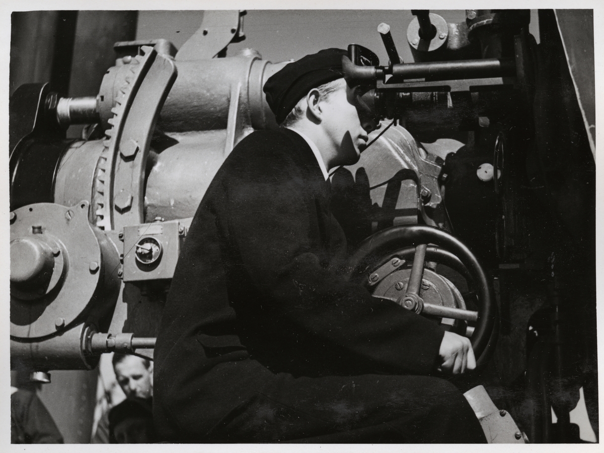 En sjöman tittar genom kikarsikte av en kanon från tung skeppsartilleri. Med den ena handen håller han på att snurra en vev för att rikta kanonen. Bakom honom på vänster sidan ser man tillslutsmekanismen.