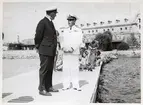 Bilden visar viceamiral Fabian Tamm i samtal Marindisktrikt Syds chef viceamiral Gösta Ehrensvärd. Dem står på en brygga framför bastionen Kungshall på Stumholmen, Karlskrona. I bakgrunden syns soldater sittande på bryggan som håller på att klä om sig före eller efter simning.
