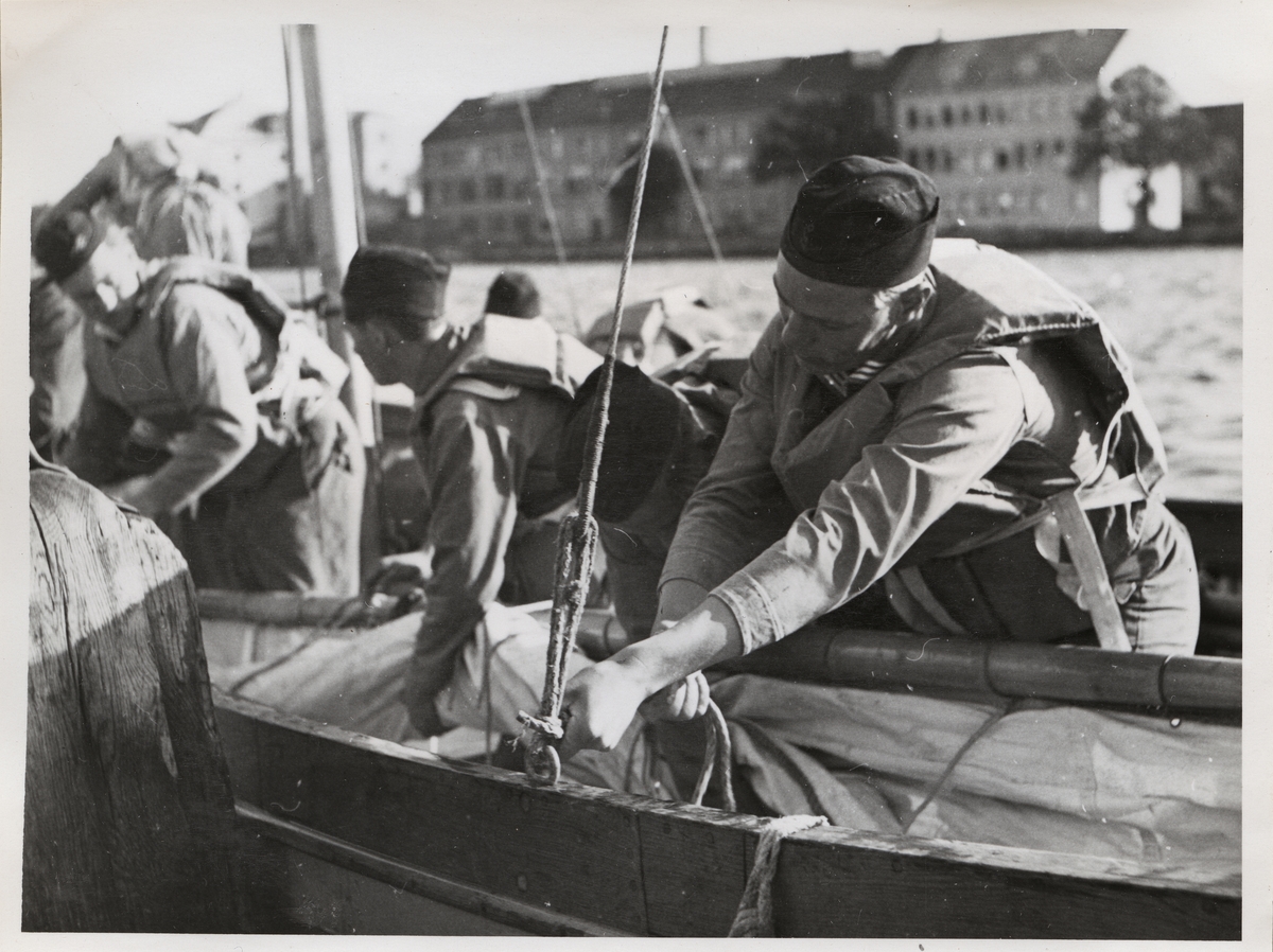 På bilden ses tre unga män täcka över en båt efter segling. Fokus i bilden ligger på den man som befinner sig i förgrunden till höger. Utöver sin uniform bär han en flytväst, liksom övriga i bilden. Männen som står till vänster i bilden befinner sig ur fokus. I bakgrunden ses beklädnadsverkstaden och beklädnadsförrådet på Stumholmen.