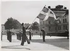 Fotografiet visar en militär ceremoni på kasernområdet i Karlskrona. Centralt i bilden befinner sig en fanbärare som håller en tretungad örlogsflagga framför sig. Han bär marinens mässdräkt komplett med handskar, skärmmössa samt sabel på den vänstra höften. Strax bakom honom till höger syns medlemmar ur marinens musikkår med sina instrument, bland annat en man med en trombon och en puka skymtar också fram bakom fanbäraren. Totalt syns nio av medlemmarna i musikkåren där de står i enskild ställning. Framför dessa står flaggtrumslagaren, till höger om fanbäraren. 
Till höger om fanbäraren ses också fem högre officierare som står spritt över kaserngården: två i mellangrunden och tre i bakgrunden. Samtliga gör honnör. 
I bakgrunden ses också yngre rekryter stå uppställda - samtliga längsmed kasernbyggnaden.