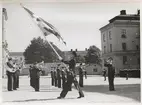 Fotografiet visar en fanbärare på kaserngården vid kasern Jarramas och kasern Najaden. Han är placerad centralt i bilden och är vänd mot vänster. Fanan han håller i är en tretungad örlogsflagga och fanstången är krönt med kung Gustav V:s monogram samt krona. Fanbäraren är själv klädd i uniform med skärmmössa, vita handskar samt en sabel vi höften. Bakom honom, till vänster ses medlemmar ur  Flottans musikkår i Karlskrona spelandes bland annat tuba och trombon. Strax till höger om fanbäraren ses också flaggtrumslagaren. Till höger i bild ses också fyra officerare utspridda på gården; samtliga gör honnör. I bakgrunden ses en av kasernbyggnaderna och längsmed väggen står rekryter.