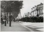 Fotografiet avbildar en parad som marscherar på Vallgatan i Karlskrona. Marschledet ses till höger i bild och sträcker sig från mellangrund till bakgrund och består huvudsakligen av unga sjömän. Jämsides med dem går också högre befäl, varav samtliga sex stycken hälsar sina betraktande kollegor med honnör. 
Till vänster i bildens förgrund finns två av dessa: en kommendör som även han gör honnör, samt en kommendörkapten som står snett bakom den förstnämnda. Bakom dessa två ses också medlemmar ur marinens musikkår; bland annat flaggtrumslagaren. I bakgrunden ses en rekryt stå vid en skara med barn som tittar på paraden. I övrigt ses i bakgrunden bebyggelse, träd och delvis Amiralitetsklockstapeln.