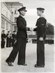 Leende officerare skakar hand med värnpliktig ung man under cermoni och medaljutdelning. I bakgrunden syns övriga befäl och värnpliktiga på rad samt en del av en byggnad.