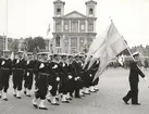 Flottans militärer under march på stortorget. Ett högre befäl tar täten med flaggan över axeln. I bakgrunden syns Fredrikskyrkan.