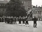 Matroser och sjöbefäl marcherar i kolonn. Längst fram bär en av matroserna en flagga och i täten går ett högre befäl. I bakgrunden syns husfasaderna omkring stortorget.