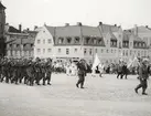 Militärer marcherar i kolonn på torget. I bakgrunden syns publik men också andra deltagande i svensk folkdräkt.