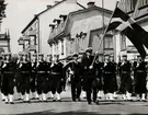 Matroser i traditionsenlig uniform marcherar nedför gata med sjöbefäl i täten, en håller en svensk flagga.