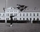 En uniformsklädd fänrik står på Sparres kaserngård med Sveriges flagga i händerna. I bakgrunden syns uppradade flottister framför matinrättningsbyggnaden.