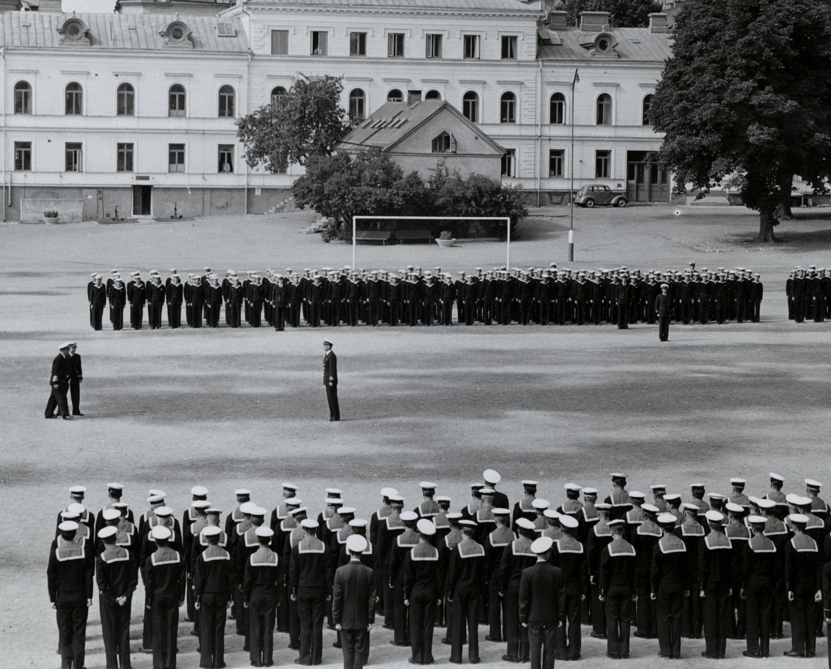 Flottister uppställda på Sparre kaserngård i två block. Mellan de två blocken syns tre militärer av högre rang. I bakgrunden syns bataljon Sparres kanslibyggnad från omkring 1818.