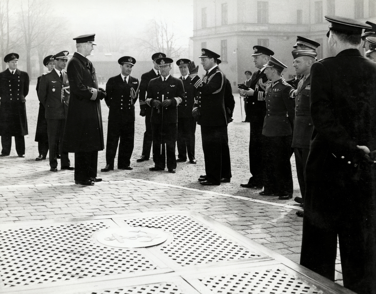 Konteramiral Samuelson och kommendörkapten Lind af Hageby står utomhus på en öppen yta tillsammans med en grupp höga militärer. Samtliga är uniformsklädda.