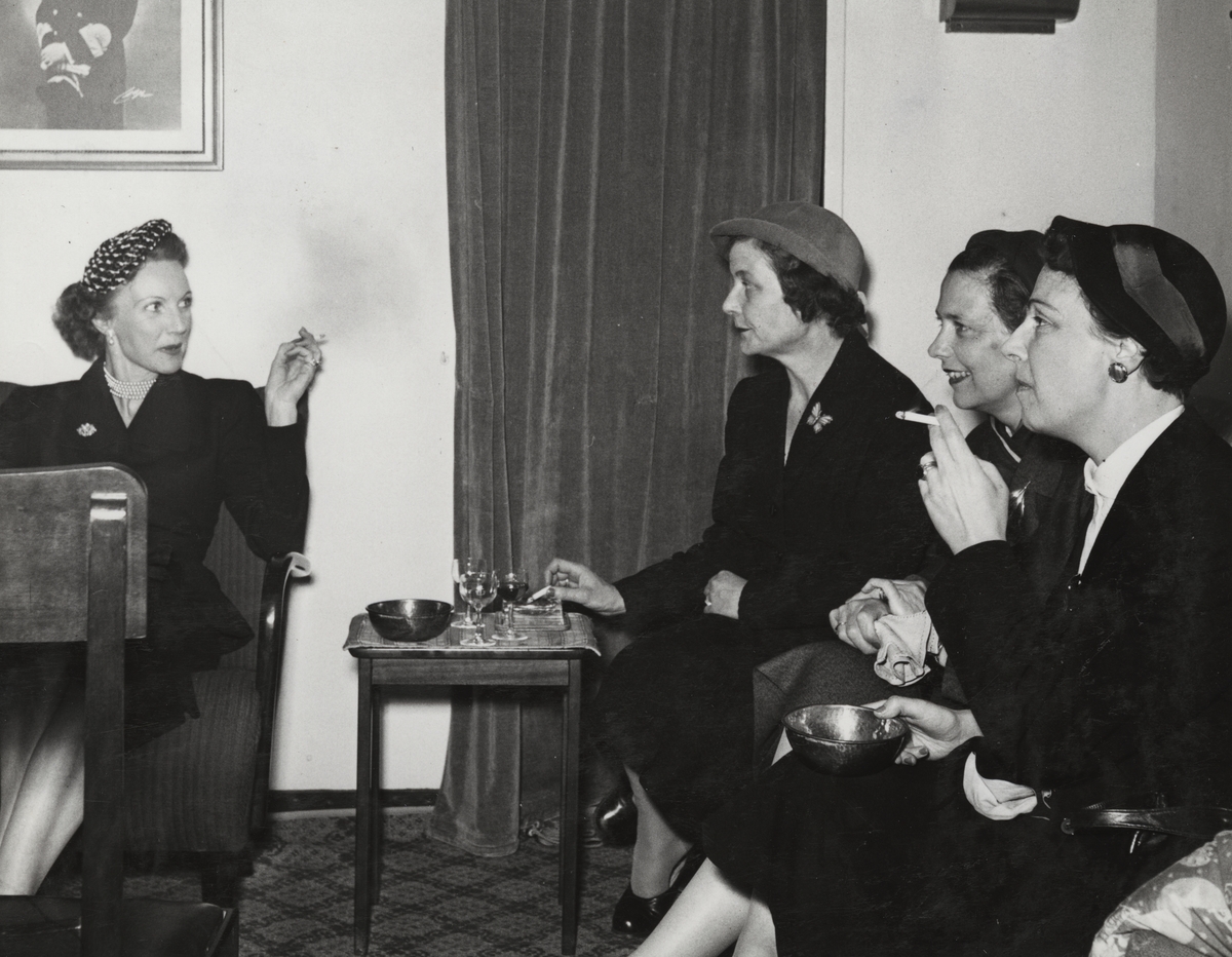 Fyra dräktklädda kvinnor i hatt sitter i ett rum och röker.