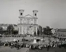 Stortorget i Karlskrona Med Fredrikskyrkan centralt i bild. Det är Svenska flaggans dag som firas med en parad av människor på torget. Folk från marinen står även uppradade och är delaktiga. Runt statyn av Karl XI har Sveriges flagga hissats på flaggstängerna. Ljudluckorna till klocktornen är öppna och troligtvis ringer det i dem när bilden tas.