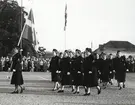 Uniformsklädda lottor marscherar över Stortorget i Karlskrona under ledning av en lotta som håller i en svensk flagga. I bakgrunden står en folksamling och tittar på marschen.