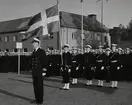 En grupp sjömän står givakt på stortorget. En fänrik i framledet håller i en stor, svensk tretungad flagga. Framför honom står en kapten. I bakgrunden syns det gamla posthuset på adressen Stortorget 3.