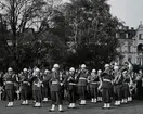 En militär musikkår framför musik. Musikerna är klädda i uniform. I bakgrunden skymtar en folksamling.