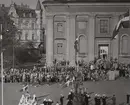 En folksamling har samlats utanför Fredrikskyrkan på Stortorget i Karlskrona. Till vänster framför folksamlingen står en militärklädd musikkår och spelar musik. I bild syns även flottister som gör honör.