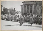 Uniformsklädda lottor står uppställda på stortorget utanför Trefaldighetskyrkan i Karlskrona. Lottan längst fram håller i en svensk flagga. I bakgrunden syns åskådare stå på Trefaldighetskyrkans trappa.