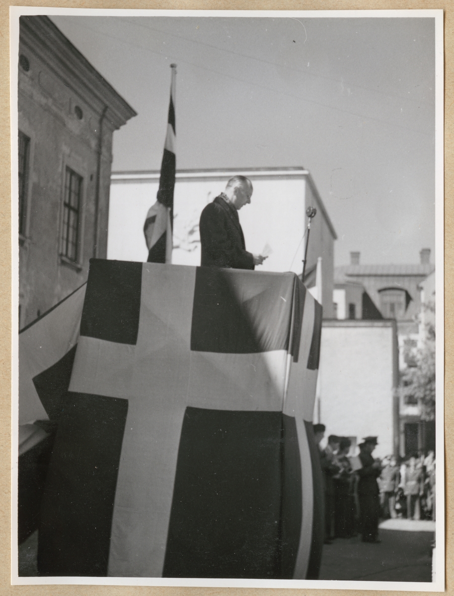 En man står i ett podium inklätt med svenska flaggan på stortorget i Karlskrona och pratar i en mikrofon. I händerna håller han ett papper. I bakgrunden skymtar rådhuset och konserthusteatern.