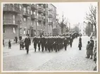 Uniformsklädda flottister marscherar i ett långt led längs med Drottninggatan i Karlskrona. Civila åskådare står utspridda på den stenbelagda gatan. Gatan kantas av flerbostadshus med utstickande balkonger.