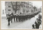 Uniformsklädda flottister marscherar längs med Drottningsgatan i Karlskrona. Längs med den stenbelagda gatan står även civila åskådare. Gatan kantas av flerbostadshus med utstickande balkonger. På gatan syns även ett parkerat motorfordon.