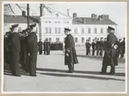 Uniformsklädda flottister står uppställda på Sparres kaserngård. Framför dem står två militärer av högre rang, den ena konteramiral. I bakgrunden syns Sparres matinrättning.