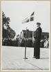 En uniformsklädd man i glasögon står framför ett stativ med en mikrofon på Stortorget i Karlskrona. I vinden svajar två svenska flaggor. I bakgrunden syns flera uniformsklädda militärer samt civila stå framför ett hus.