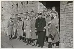 En uniformsklädd kommendör poserar framför kameran med en grupp uniformsklädda lottor vid en mur på Kungsholmen.