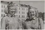 Två lottor ler och poserar mot kameran, iklädda i mössor och uniformer. I bakgrunden syns Östra Kasernen på Gräsvik.