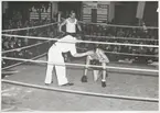 Två boxaren och en domare står i en boxningsring. Domaren står lutad över den ena boxaren, som står på ett knä. Den andra boxaren står i ena hörnan. Runt omkring finns åskådare.