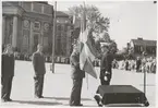 En uniformsklädd militär står på stortorget i Karlskrona och har lämnat över en svensk flagga till en man klädd i kostym. Bakom den civilklädda mannen står ytterligare två kostymklädda män. I händerna har de varsin hatt. Bredvid militären står ett litet bord med en duk på samt en mikrofon. I bakgrunden syns delar av Trefaldighetskyrkan samt en folksamling som bevittnar händelsen.