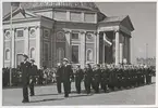 Flottister på marsch genom stortorget. Framför dem går en kapten. I bakgrunden syns civila åskådare uppradade framför Trefaldighetskyrkan.