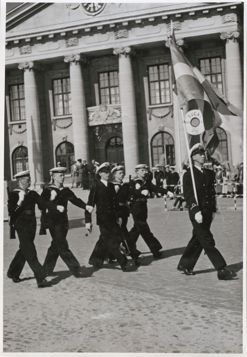 Uniformsklädda flottister väpnade med skjutvapen marscherar framför Karlskrona rådhus. Längst fram går en fänrik som håller i en svensk flagga. Rådhusets fronton bärs upp av joniska kolonner och fasaden smyckas med festonger.