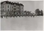 Uniformsklädda lottor står uppställa på en grusyta utanför kanslihuset på Gräsvik, uppfört 1906. Flera av dem håller i svenska flaggor.