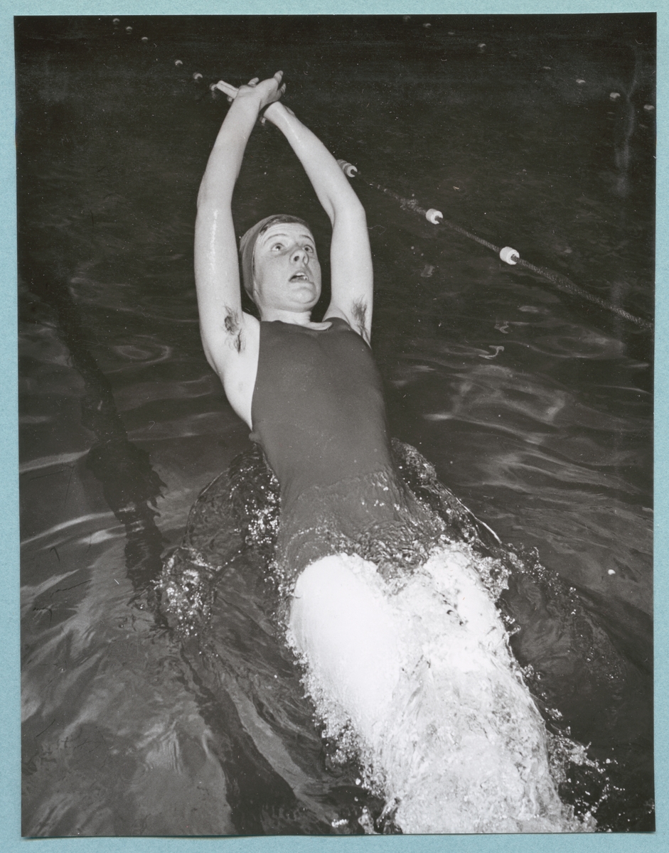 En kvinnlig simmare, i baddräkt och badmössa, simmar på rygg i bassängen tillhörande Sparres simhall. Fotot är daterat till 15-3-1952.