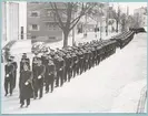 Uniformsklädda flottister marscherar längs med Drottninggatan i Karlskrona. I täten går en fänrik. Gatan kantas av bebyggelse, bl.a. i form av ett lägenhetshus med utskjutande balkonger. Fotot är daterat till 17-3-1952.