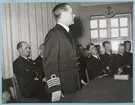 En kommendörkapten av första graden står vid ett bord och talar. Runt bordet sitter flera militärer, bl.a. kaptener, löjtnanter och fänrikar. Samtliga är klädda i uniform.