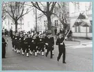 En grupp uniformsklädda flottister marscherar på Drottninggatan. Längst fram går en fänrik som håller i en flagga. Längs med gatan syns både civila och militärer som tittar på marschen. I bakgrunden syns bataljon Sparre.