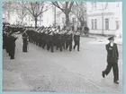 Flera grupper av uniformsklädda flottister marscherar längs med Drottningsgatan, förbi bataljon Sparre. En liten bit framför den första gruppen går en kapten. På gatan står en militär musikkår och spelar musik. På andra sidan gatan, utanför bataljon Sparre, står ytterligare militärer och tittar på.