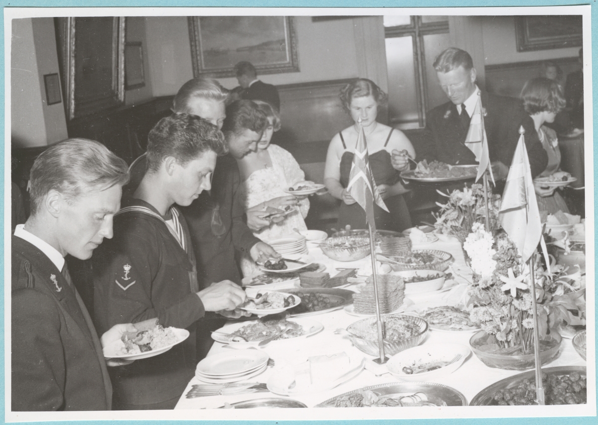 Från dansen och supén på Sjöofficersmässen. Ett bord står uppdukat med mat, blommor och den danska, svenska och norska flaggan. En grupp flottister samt civila kvinnor i klänningar lägger upp mat åt sig.